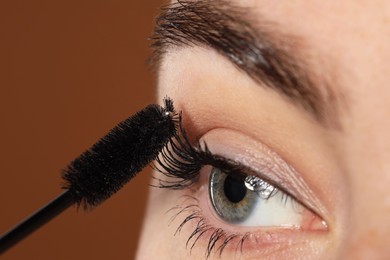 Photo of Woman applying mascara onto her eyelashes on brown background, closeup