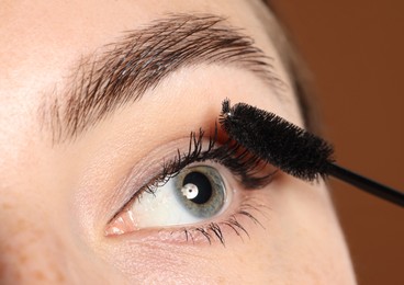 Photo of Woman applying mascara onto her eyelashes on brown background, closeup