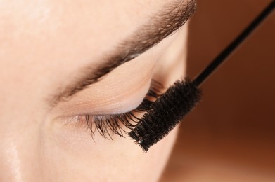 Photo of Woman applying mascara onto her eyelashes on brown background, closeup