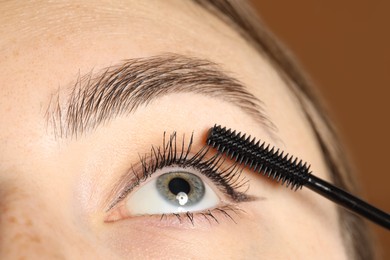 Photo of Woman applying mascara onto her eyelashes on brown background, closeup