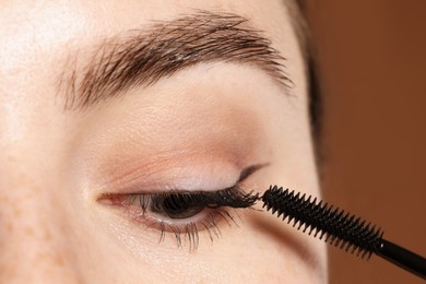 Photo of Woman applying mascara onto her eyelashes on brown background, closeup