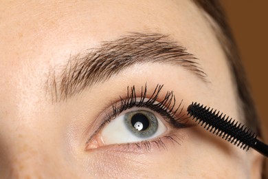 Photo of Woman applying mascara onto her eyelashes on brown background, closeup
