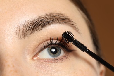 Photo of Woman applying mascara onto her eyelashes on brown background, closeup