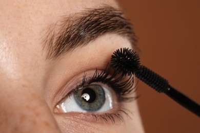 Photo of Woman applying mascara onto her eyelashes on brown background, closeup