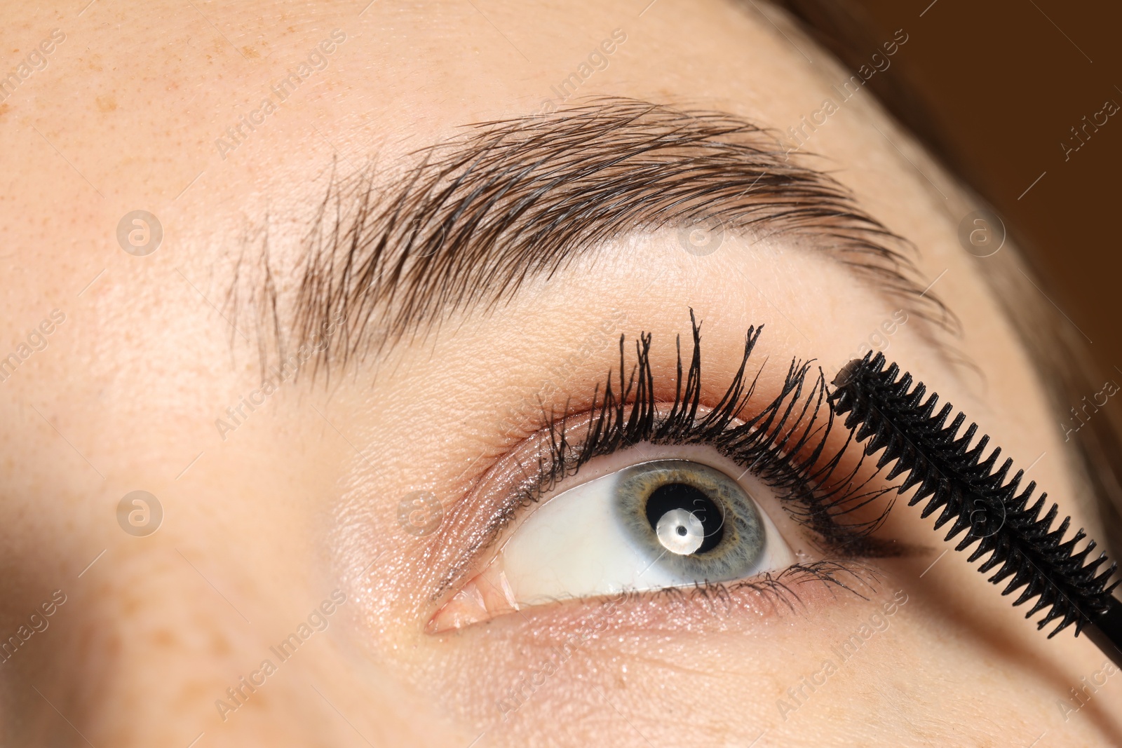 Photo of Woman applying mascara onto her eyelashes on brown background, closeup