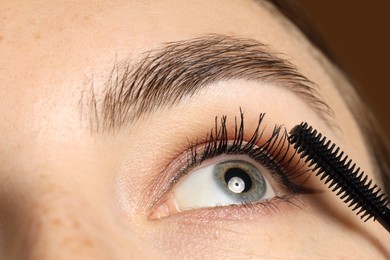 Photo of Woman applying mascara onto her eyelashes on brown background, closeup