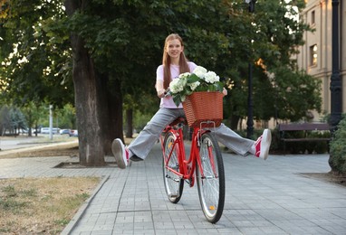 Photo of Beautiful teenage girl riding bicycle with basket of flowers on city street