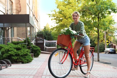 Photo of Beautiful woman on bicycle with basket of flowers on city street, space for text