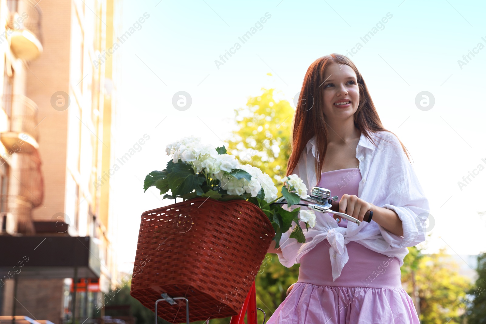 Beautiful teenage girl near bicycle with basket of flowers on city street, low angle view Photo of Beautiful teenage girl near bicycle with basket of flowers on city street, low angle view