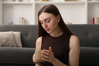 Photo of Beautiful woman with cup of drink near sofa at home