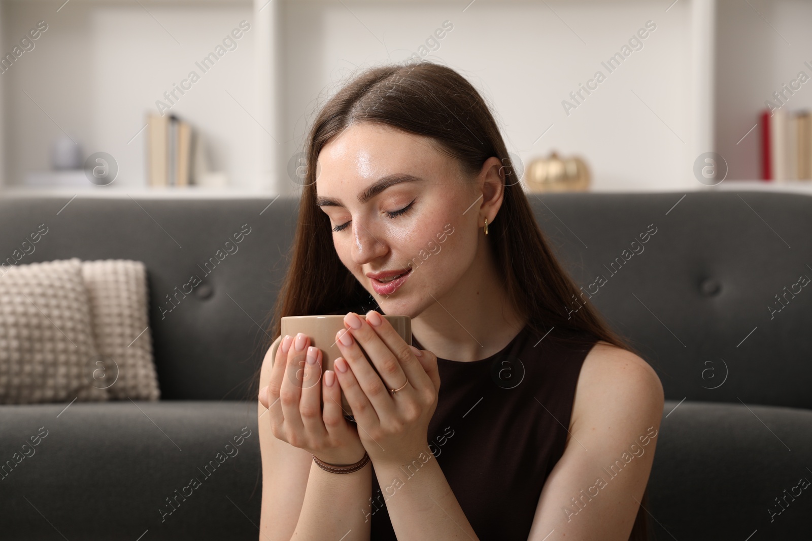 Photo of Beautiful woman with cup of drink near sofa at home