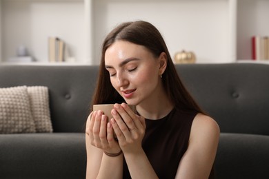 Photo of Beautiful woman with cup of drink near sofa at home