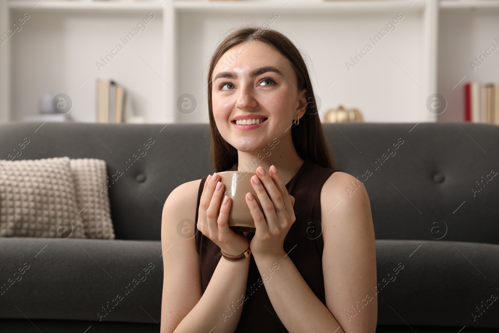 Photo of Beautiful woman with cup of drink near sofa at home