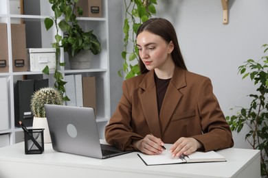 Photo of Woman taking notes while working with laptop at desk in office