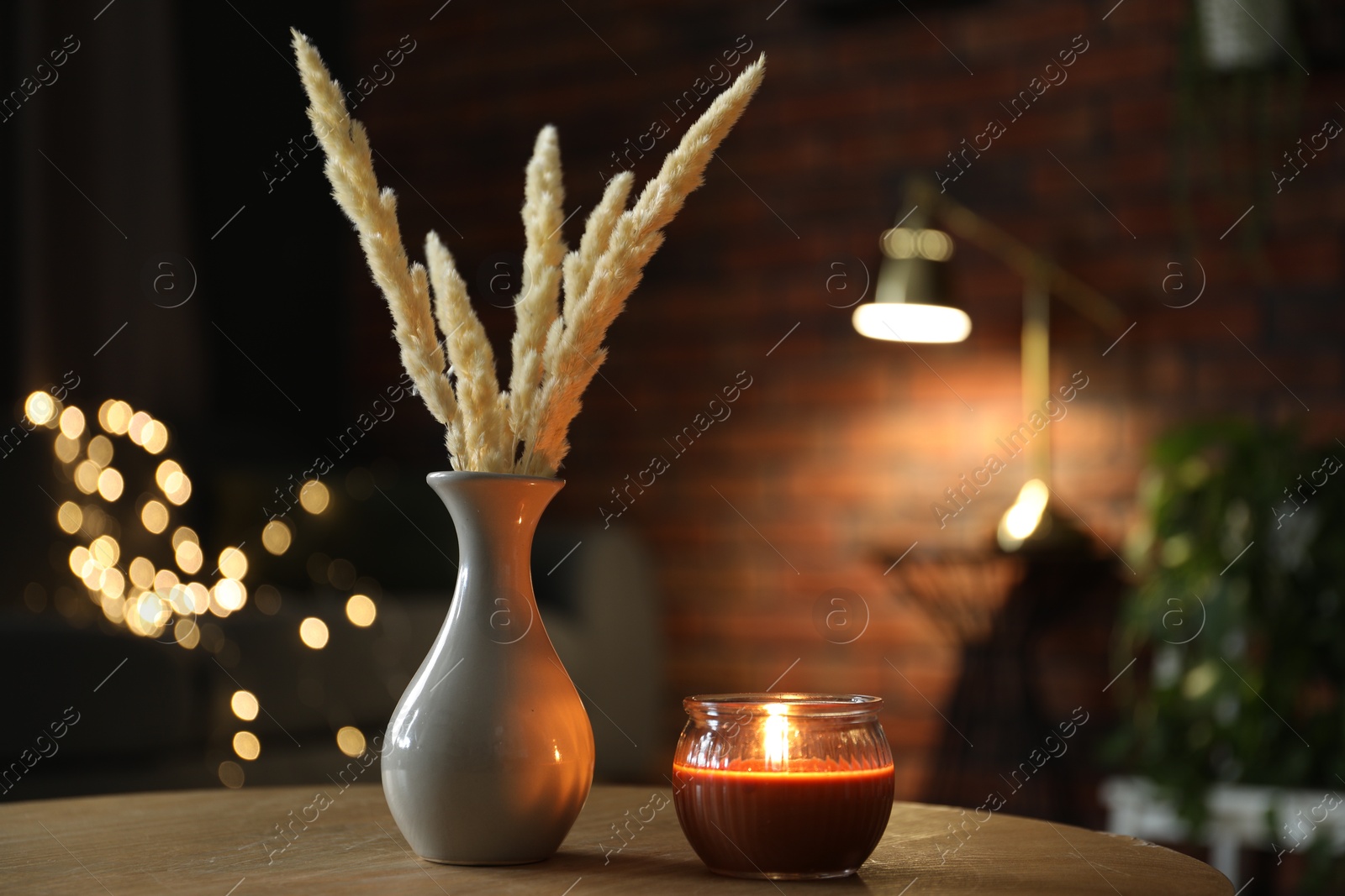 Photo of Burning candle and vase with spikelets on wooden table against blurred lights. Space for text