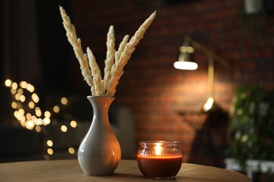Photo of Burning candle and vase with spikelets on wooden table against blurred lights. Space for text