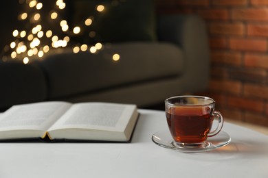 Photo of Cup of tea and book on white table against blurred lights