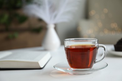 Photo of Cup of tea and book on white table against blurred lights, closeup