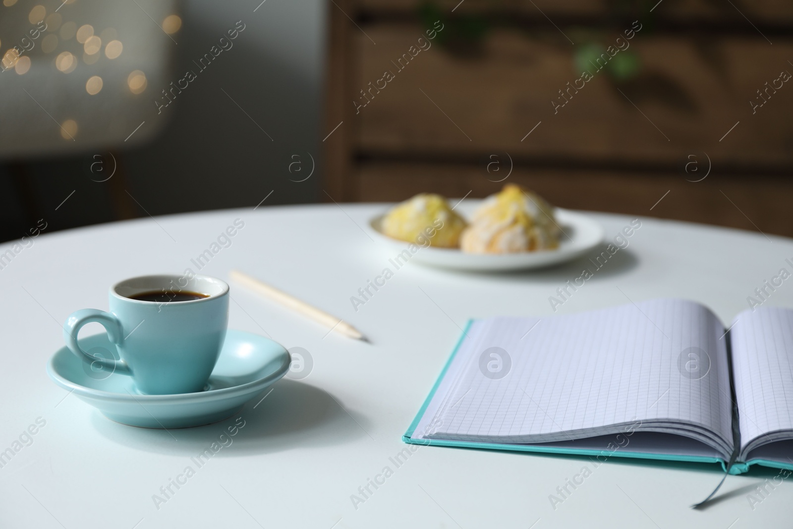 Photo of Notebook, cup of coffee and cookies on white table against blurred lights