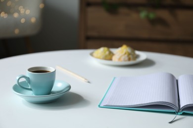 Photo of Notebook, cup of coffee and cookies on white table against blurred lights