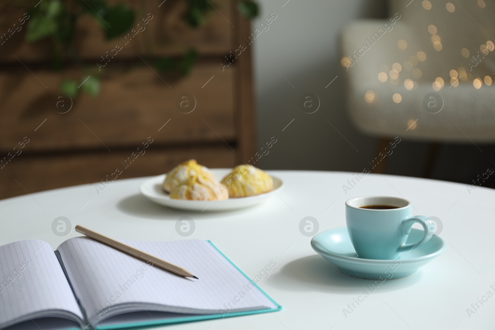 Photo of Notebook with pencil, cup of coffee and cookies on white table against blurred lights