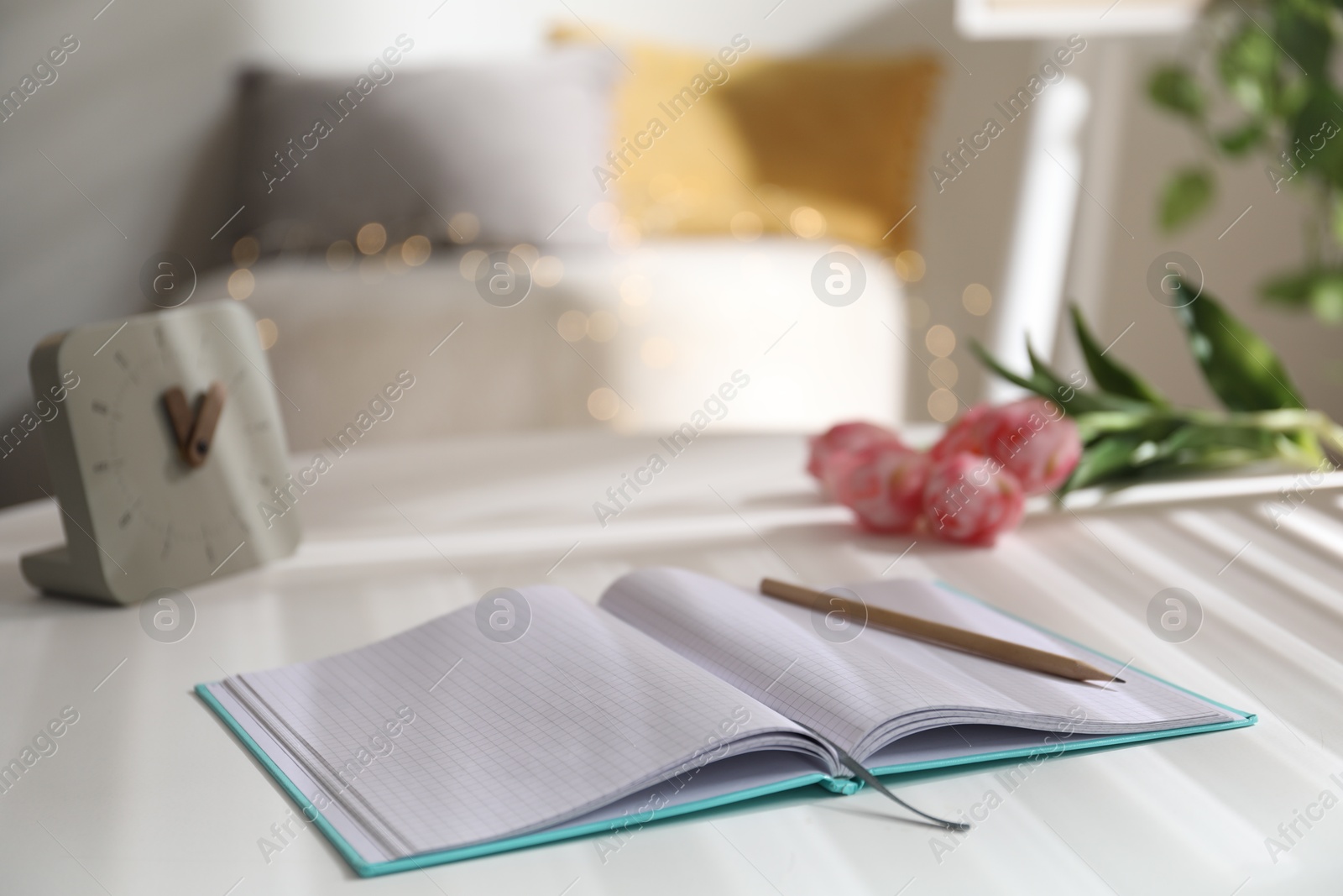 Photo of Notebook with pencil, alarm clock and tulips on white table against blurred lights