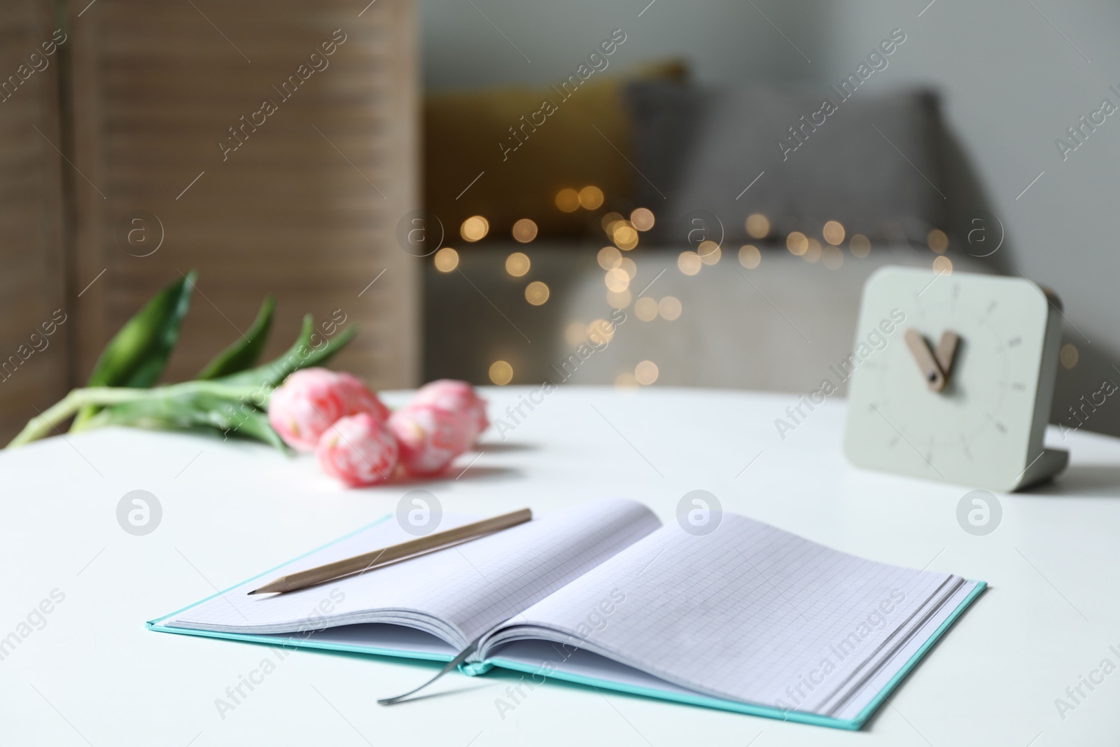 Photo of Notebook with pencil, alarm clock and tulips on white table against blurred lights