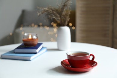 Photo of Cup of tea, books and burning candle on white table against blurred lights