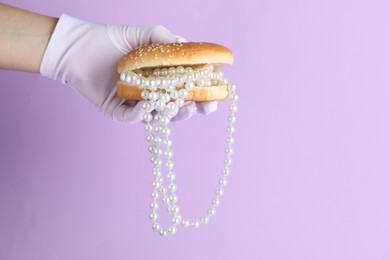 Photo of Woman holding burger with pearl necklace on lilac background, closeup
