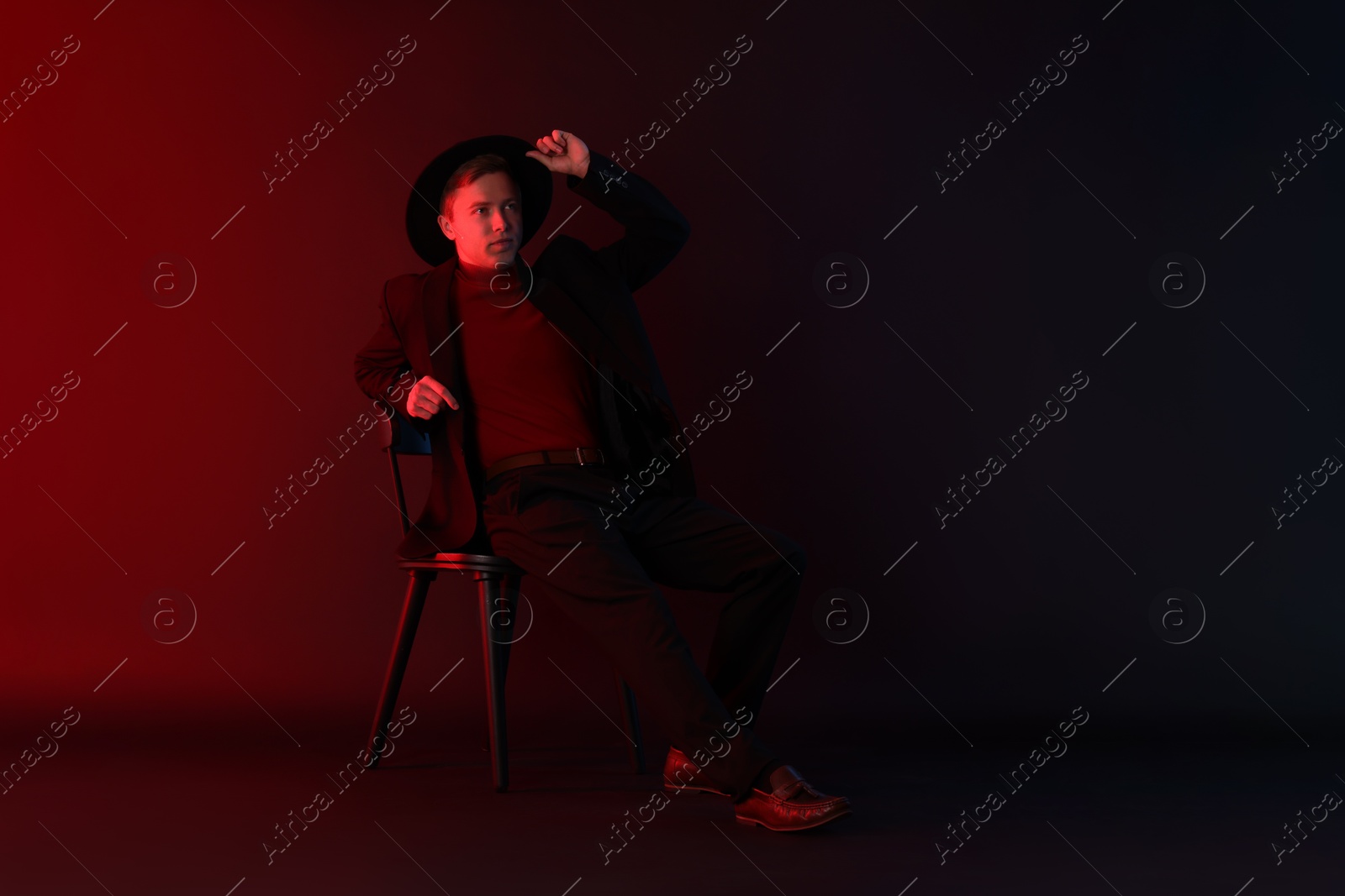 Handsome man wearing suit and hat sitting on chair in neon lights against dark background Photo of Handsome man wearing suit and hat sitting on chair in neon lights against dark background