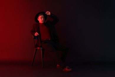 Photo of Handsome man wearing suit and hat sitting on chair in neon lights against dark background
