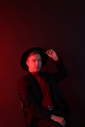 Photo of Handsome man wearing suit and hat sitting on chair in neon lights against dark background