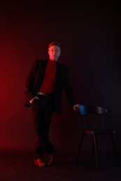 Photo of Handsome man wearing suit standing near chair in neon lights against dark background