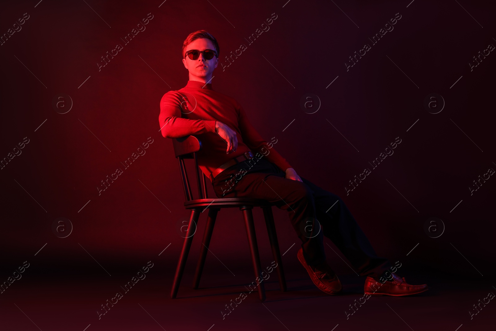 Photo of Handsome man wearing sunglasses sitting on chair in neon lights against dark background