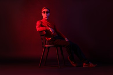 Photo of Handsome man wearing sunglasses sitting on chair in neon lights against dark background