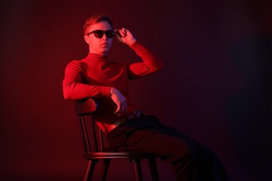 Photo of Handsome man wearing sunglasses sitting on chair in neon lights against dark background