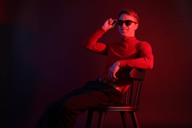 Photo of Smiling man wearing sunglasses sitting on chair in neon lights against dark background