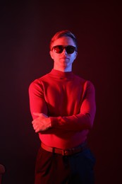 Photo of Portrait of handsome man wearing sunglasses with crossed arms in neon lights against dark background