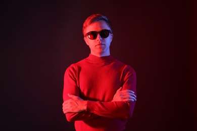 Photo of Portrait of handsome man wearing sunglasses with crossed arms in neon lights against dark background