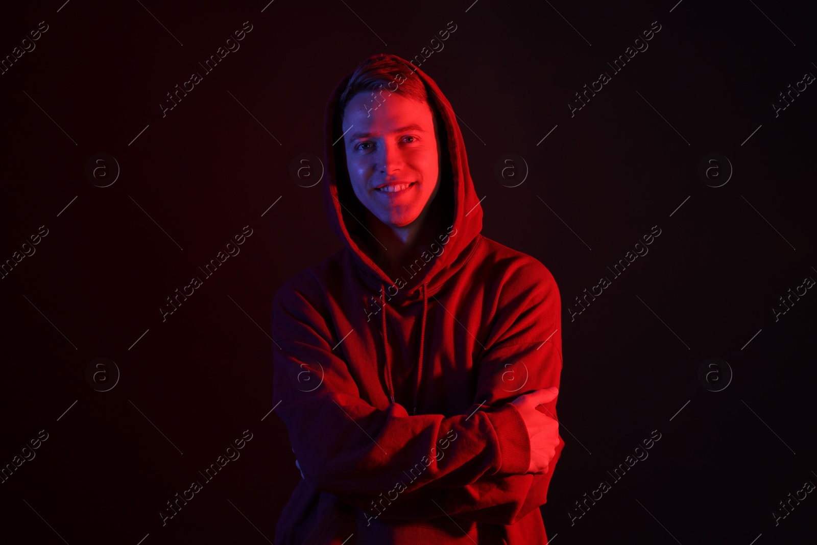 Portrait of smiling man wearing hoodie with crossed arms in neon lights against black background Photo of Portrait of smiling man wearing hoodie with crossed arms in neon lights against black background
