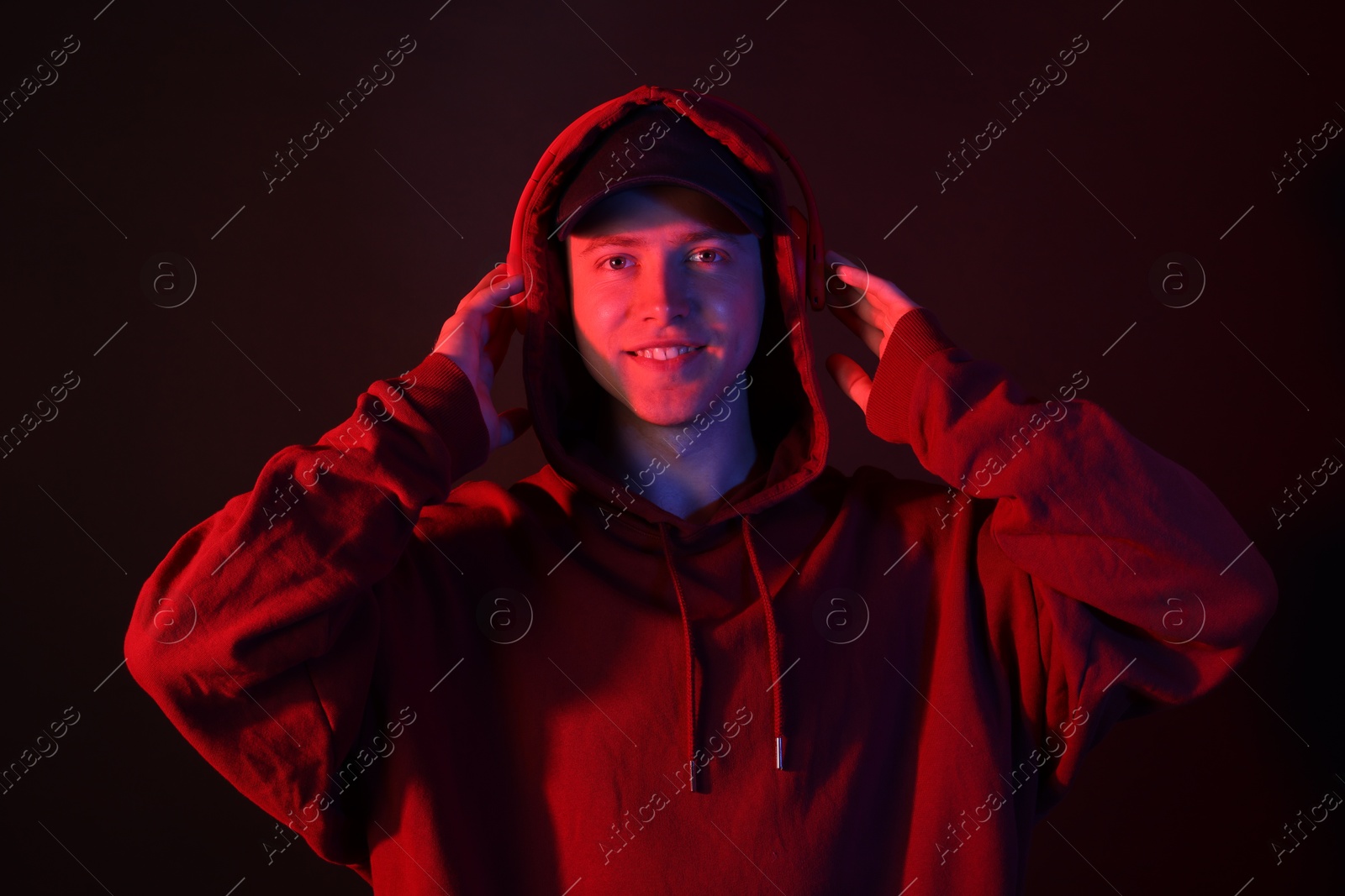 Portrait of smiling man wearing hoodie, baseball cap and headphones in neon lights against black background Photo of Portrait of smiling man wearing hoodie, baseball cap and headphones in neon lights against black background