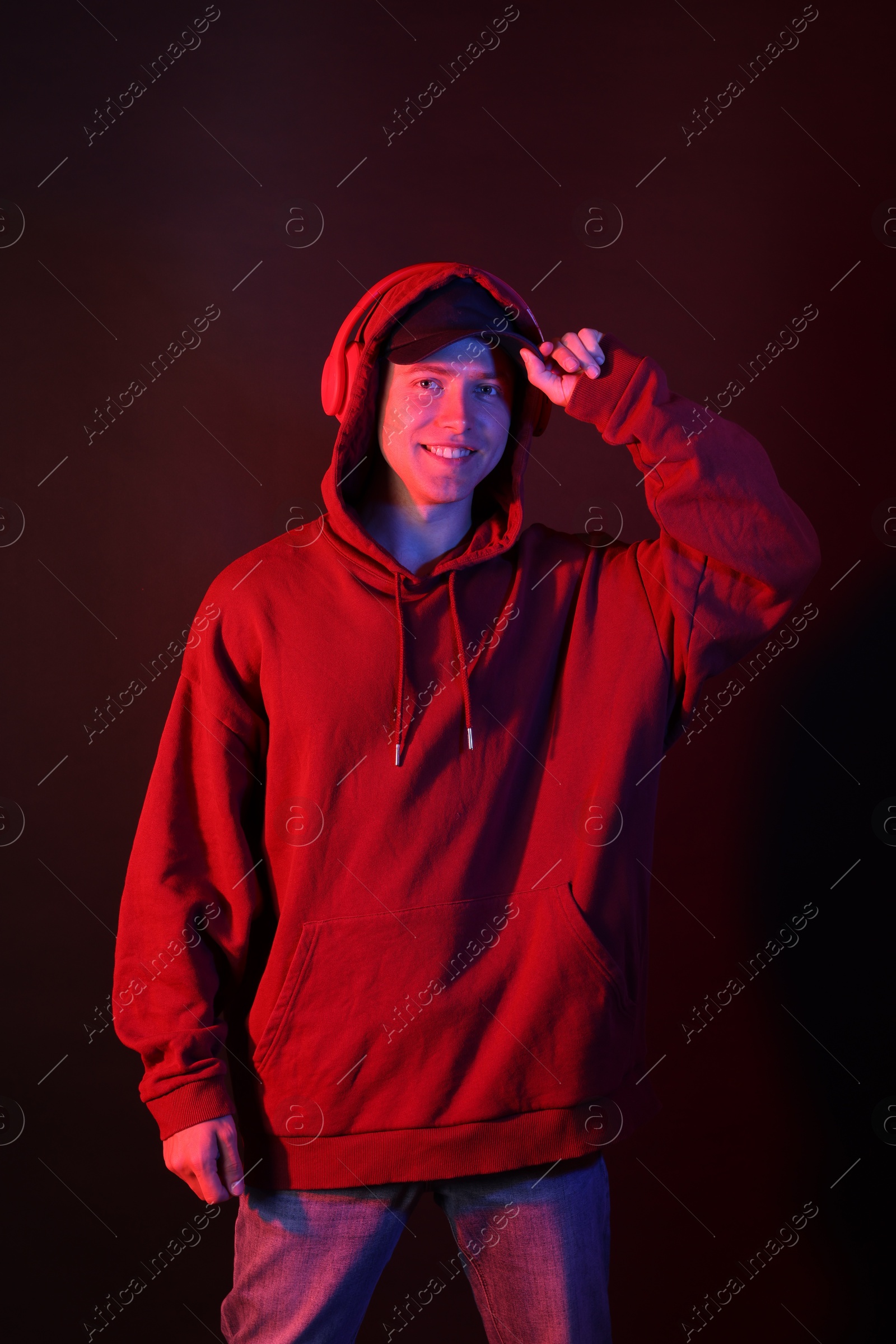 Portrait of smiling man wearing hoodie, baseball cap and headphones in neon lights against black background Photo of Portrait of smiling man wearing hoodie, baseball cap and headphones in neon lights against black background