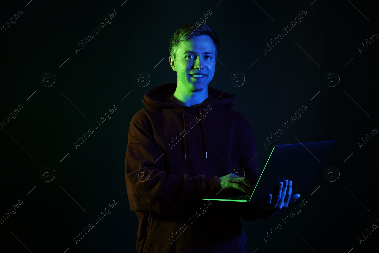 Smiling man wearing hoodie using laptop in neon lights against black background Photo of Smiling man wearing hoodie using laptop in neon lights against black background