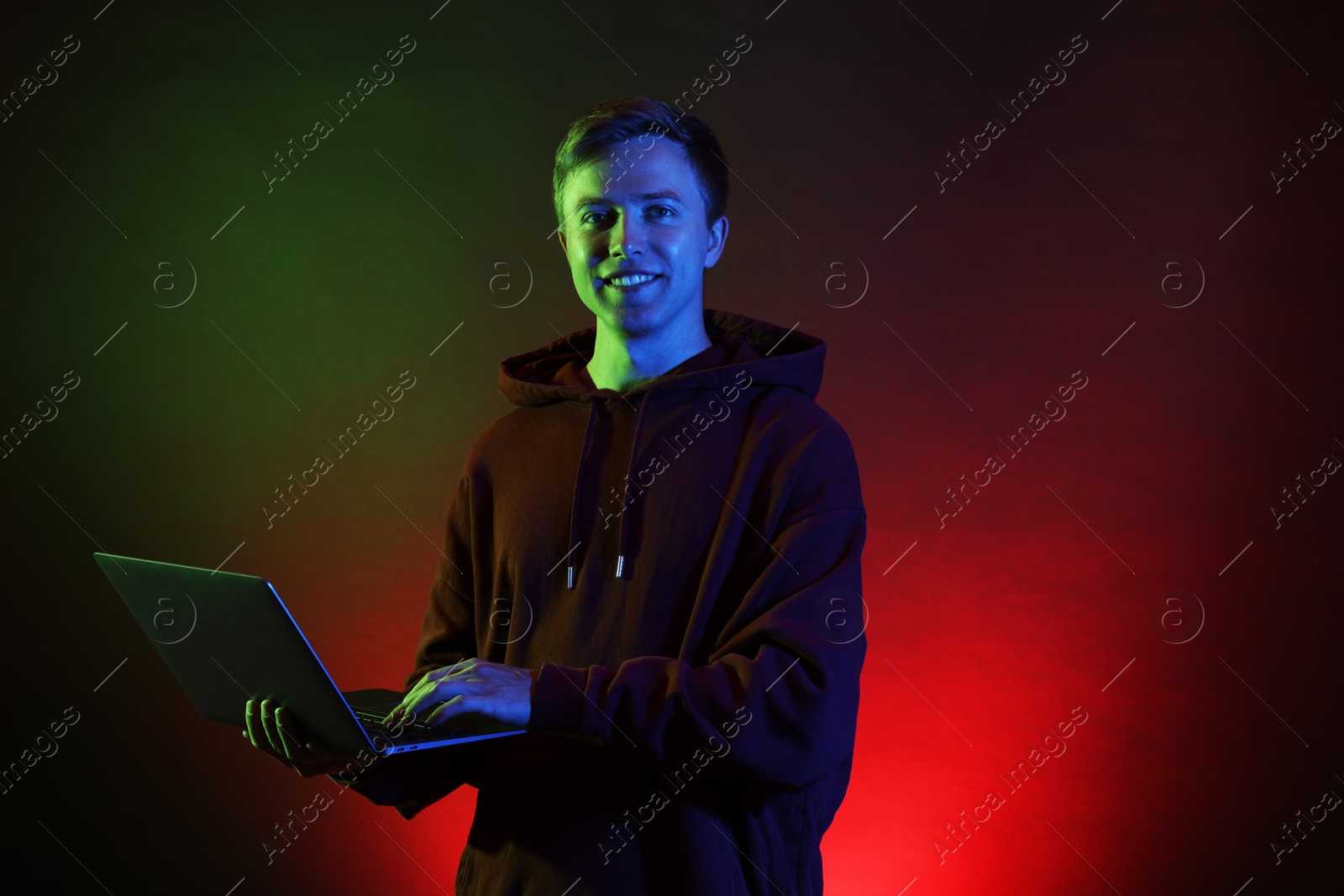 Photo of Smiling man wearing hoodie using laptop in neon lights against black background