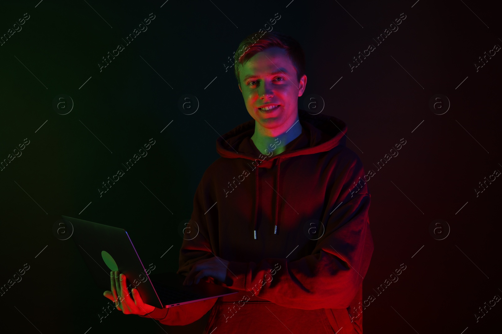 Smiling man wearing hoodie using laptop in neon lights against black background Photo of Smiling man wearing hoodie using laptop in neon lights against black background