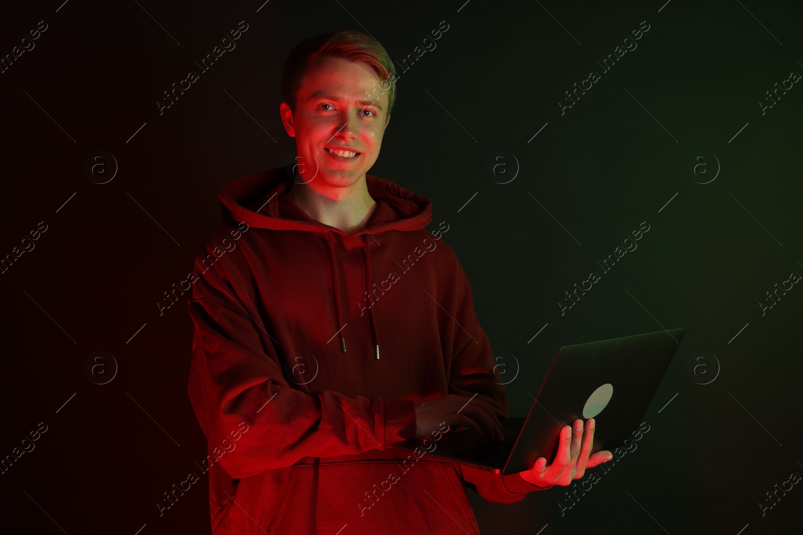 Photo of Smiling man wearing hoodie using laptop in neon lights against black background