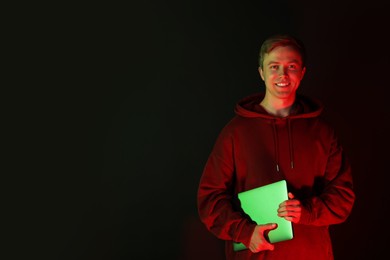 Photo of Smiling man wearing hoodie with laptop in neon lights against black background. Space for text