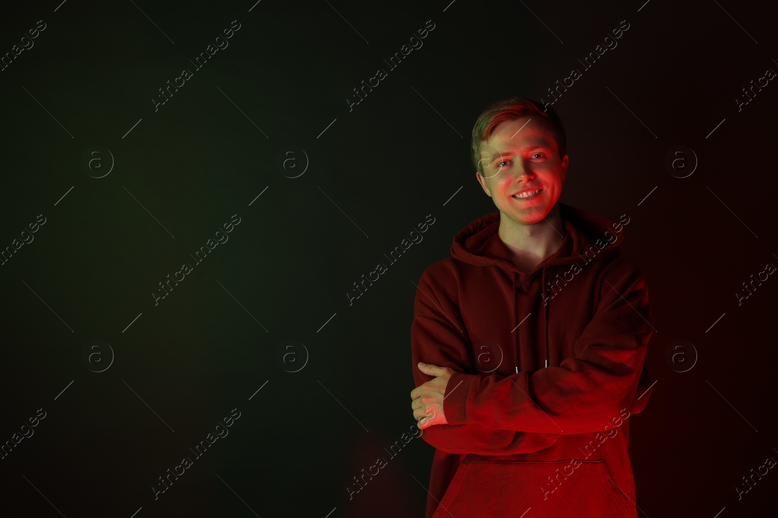 Photo of Portrait of smiling man wearing hoodie with crossed arms in neon lights against black background. Space for text
