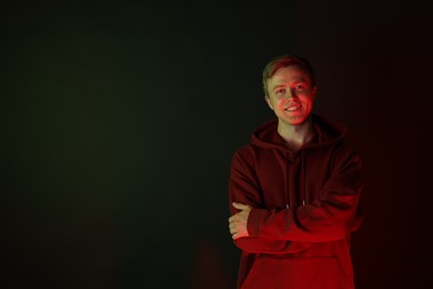 Photo of Portrait of smiling man wearing hoodie with crossed arms in neon lights against black background. Space for text