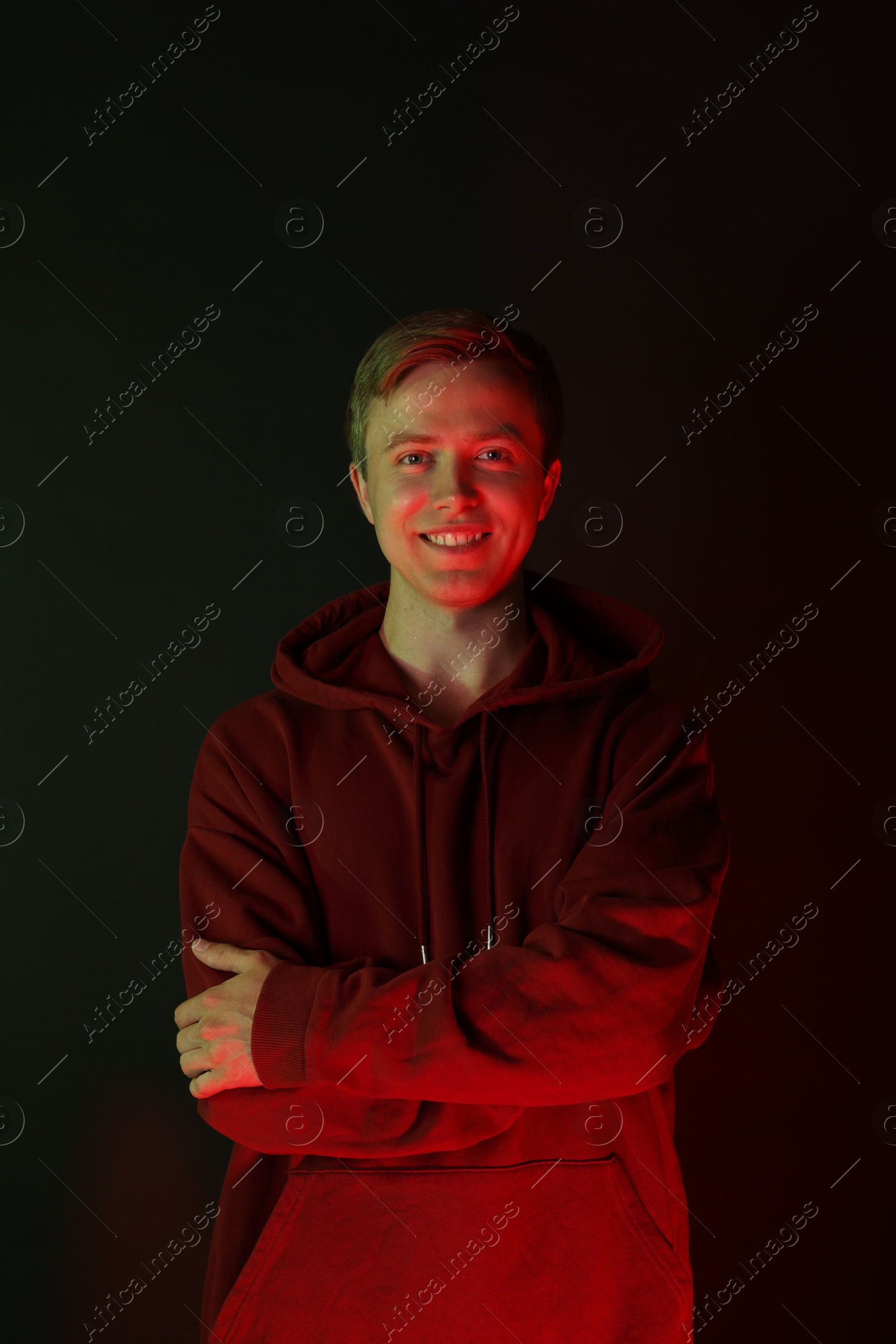 Photo of Portrait of smiling man wearing hoodie with crossed arms in neon lights against black background